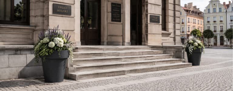 Schwarze Beton-Pflanzkübel Isabelle mit weißen und violetten Blumen, seitlich an den Stufen vor dem Rathaus auf dem Marktplatz platziert