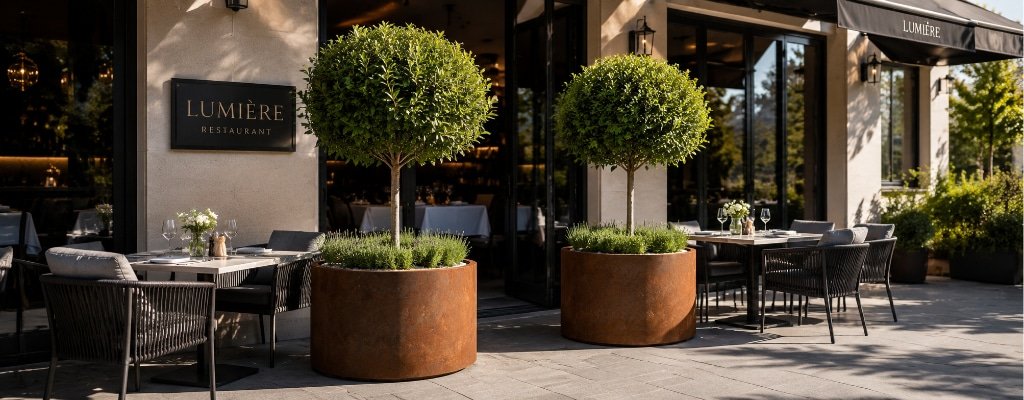 Zwei große runde Cortenstahl-Pflanzkübel mit kleinen Hochstamm-Bäumen als Sichtschutz und Raumteiler vor einer sonnigen Restaurantterrasse, die die Außentische voneinander abgrenzen