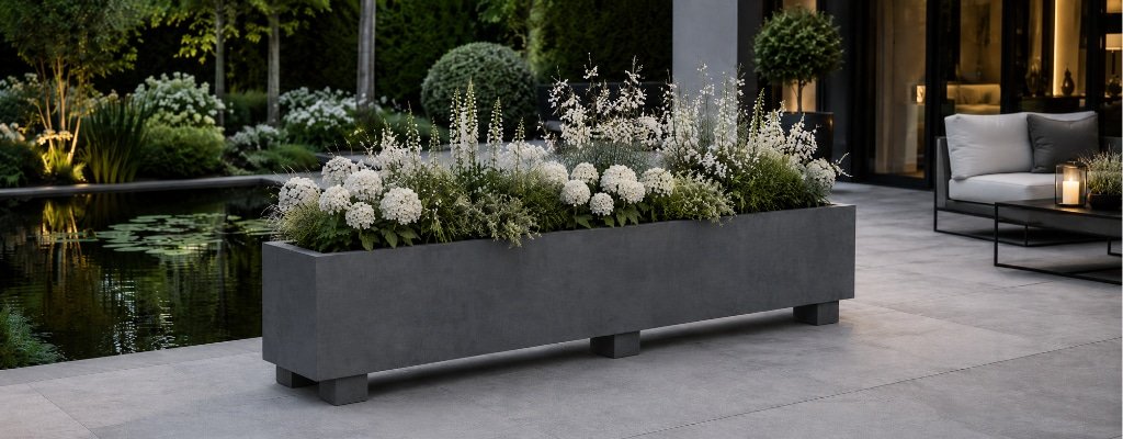 Grauer Beton-Pflanzkübel mit weißen Blumen auf der Terrasse am Gartenteich