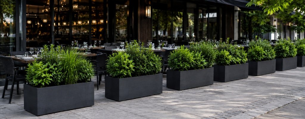Rechteckige Beton-Pflanzkübel als Raumteiler vor einer modernen Restaurantterrasse mit grüner Bepflanzung