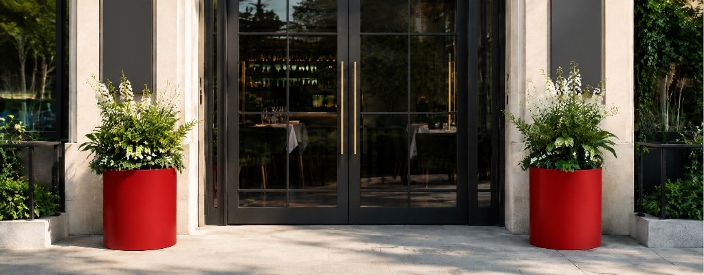 Zwei hohe rote Fiberglas-Pflanzkübel mit grün-weißer Bepflanzung vor dem Eingang einer Restaurantterrasse