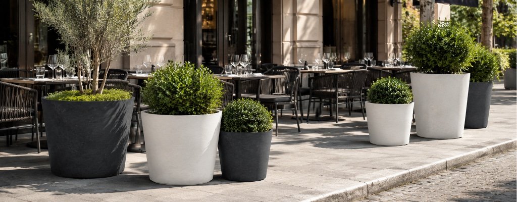 Mehrere weiße und schwarze Beton-Pflanzkübel in verschiedenen Größen vor den Außentischen einer modernen Restaurantterrasse