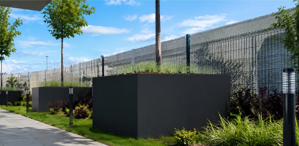 Große anthrazitfarbene Pflanzkübel aus Aluminium mit Bäumen und Ziergräsern vor einem modernen Gewerbegebäude.