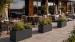 Anthrazitfarbene rechteckige Aluminium-Pflanzgefäße mit Ziergräsern, Lavendel und Stauden auf der Terrasse eines modernen Hotels.