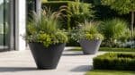Beton-Pflanzkübel im Garten, einer im Vordergrund und einer im Hintergrund, bepflanzt mit Ziergräsern und grünen Pflanzen