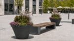 Beton-Pflanzkübel mit Ziergräsern, grünen Pflanzen und Blüten neben einer modernen Sitzbank auf einem hellen Stadtplatz