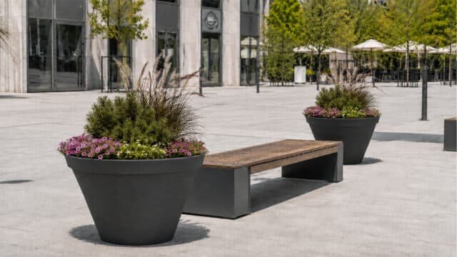 Beton-Pflanzkübel mit Ziergräsern, grünen Pflanzen und Blüten neben einer modernen Sitzbank auf einem hellen Stadtplatz