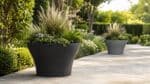 Beton-Pflanzkübel in Schwarz und Grau mit dekorativen Gräsern und grüner Bepflanzung entlang eines Gartenwegs, einer im Vordergrund und einer im Hintergrund