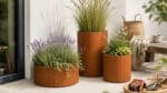 Pflanzgefäße in Cortenstahl-Optik mit Ziergräsern, Lavendel und blühenden Pflanzen auf einer hellen Terrasse mit Gartenaccessoires