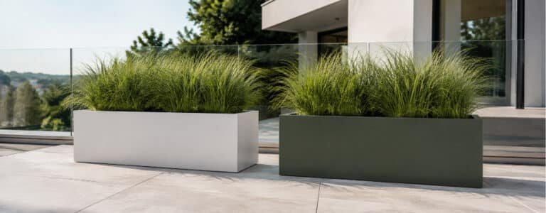 Zwei moderne rechteckige Pflanzkübel in Weiß und Dunkelgrün mit üppigen Ziergräsern auf einer sonnigen Terrasse