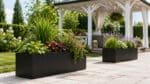 Zwei schwarze Fiberglas-Pflanzkübel mit unterschiedlichen Pflanzen auf einem Weg neben einer Gartenlaube