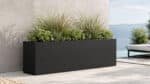 Langer schwarzer Fiberglas-Pflanzkübel mit Ziergräsern und grünen Pflanzen auf einer hellen Terrasse